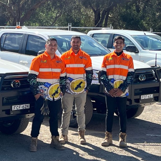 Three Diona employees in Ballarat stand in front of their vehicles, wearing hi-vis shirts adorned with Indigenous artwork.