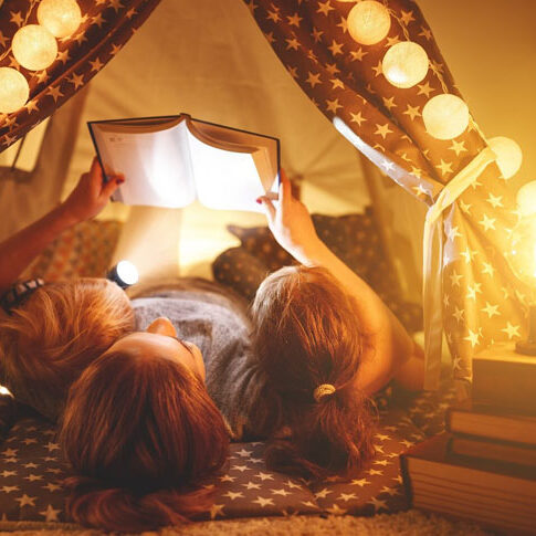 A mother reading to two children in a tent illuminated by fairy lights, symbolising the warmth and connection brought by sustainable power solutions.
