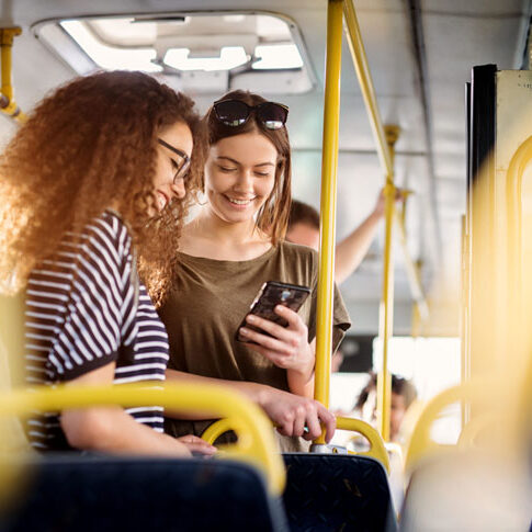 Two girls on a bus sharing a moment of connection over a smartphone, symbolising the impact of enhanced transport infrastructure.
